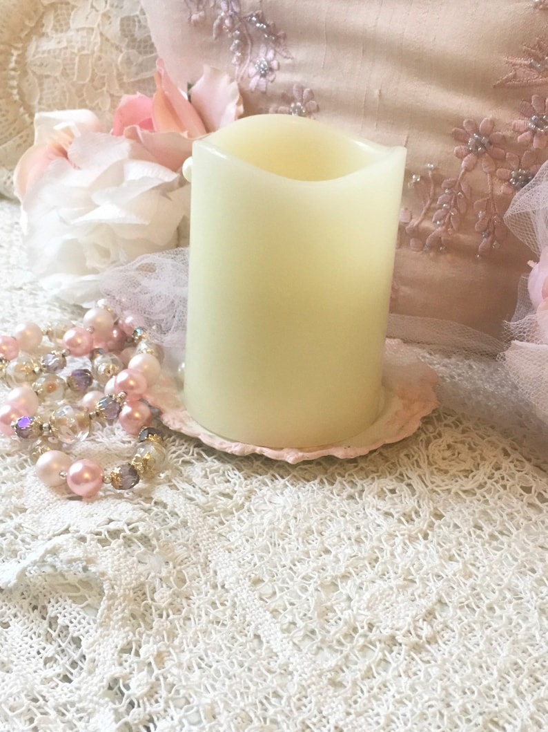 May include: A white pillar candle with a slightly curved top sits on a pink and white decorative plate. The candle is on a white lace tablecloth.