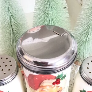 May include: A set of three vintage-style Christmas salt and pepper shakers. Each shaker features a different image of Santa Claus with a red hat and holly. The shakers have silver lids with a small hole for dispensing salt or pepper.