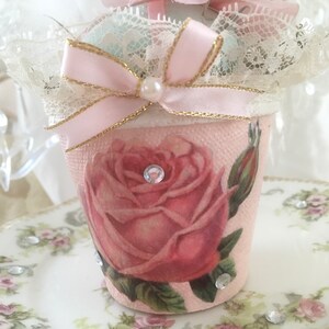Puede incluir: Un pequeño macetero de cerámica rosa con un borde de encaje blanco y una cinta rosa con detalles dorados. El macetero presenta una rosa rosa con hojas verdes y un strass en el centro. El macetero está sentado sobre un platillo blanco con un patrón floral.