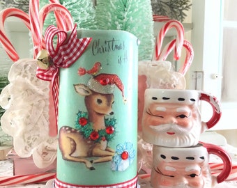 May include: A vintage-style Christmas candle holder with a green and white gingham ribbon, a red bow, and a bell. The candle holder features a deer with a red Santa hat and a red bird. The text "Christmas is Here" is printed on the candle holder. Two Santa mugs are in the background.