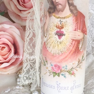 May include: A decorative candle featuring an image of Jesus with a halo, a heart, and floral accents. The candle has the text "Heiliges Herz Jesu". A lace ribbon and a rosary with pearls are also visible.