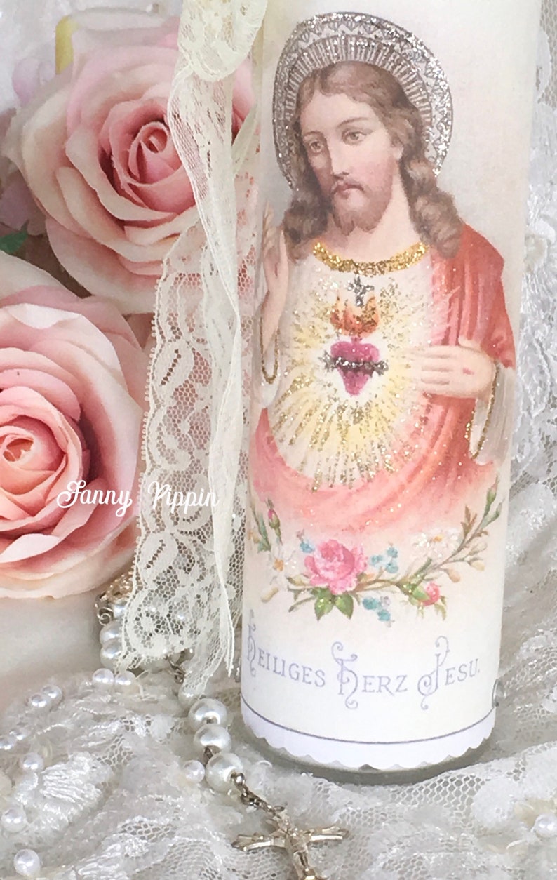 May include: A decorative candle featuring an image of Jesus with a halo, a heart, and floral accents. The candle has the text "Heiliges Herz Jesu". A lace ribbon and a rosary with pearls are also visible.