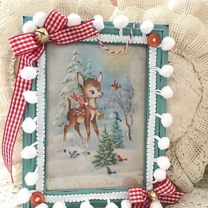 Puede incluir: Un cuadro navide&ntilde;o con una ilustraci&oacute;n vintage de un ciervo en un paisaje nevado. El marco turquesa est&aacute; adornado con pompones blancos, lazos de cuadros rojos y blancos y una peque&ntilde;a campana dorada. El marco est&aacute; adornado con encaje blanco.