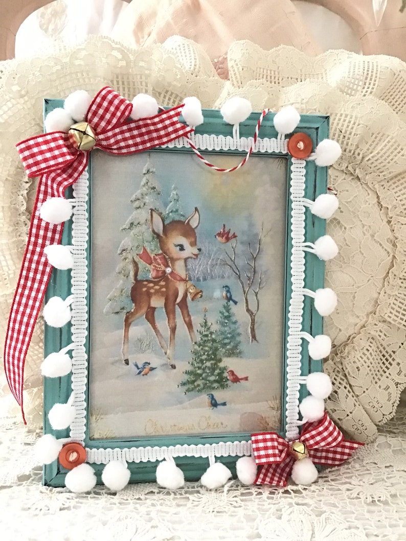 Puede incluir: Un cuadro navide&ntilde;o con una ilustraci&oacute;n vintage de un ciervo en un paisaje nevado. El marco turquesa est&aacute; adornado con pompones blancos, lazos de cuadros rojos y blancos y una peque&ntilde;a campana dorada. El marco est&aacute; adornado con encaje blanco.