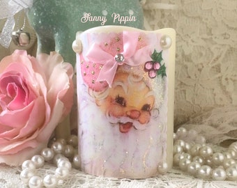May include: A white candle with a pink bow and a vintage Santa Claus image. The candle has a glittery finish and a pink and gold polka dot pattern.
