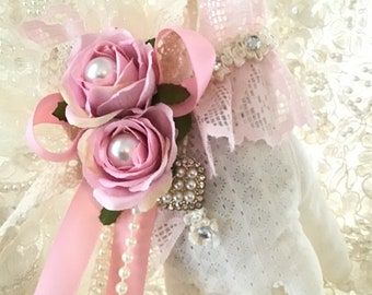 May include: A pair of white lace gloves with a pink satin ribbon and two pink roses with pearl centers. The gloves have a pink lace trim and a rhinestone embellishment.