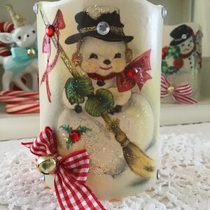 May include: A white ceramic candle holder with a vintage image of a snowman holding a broom. The snowman is wearing a black top hat with a gold band and a red bow tie. The candle holder has a red and white gingham bow with a gold bell attached.