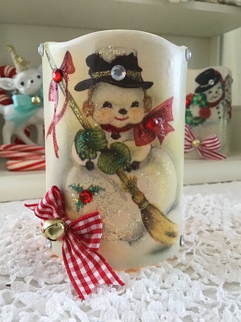 May include: A white ceramic candle holder with a vintage image of a snowman holding a broom. The snowman is wearing a black top hat with a gold band and a red bow tie. The candle holder has a red and white gingham bow with a gold bell attached.