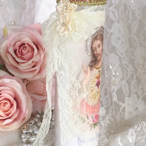 May include: A decorative candle with an image of Jesus, adorned with lace, ribbon, and floral accents. The candle features a gold and pink jeweled trim at the top and is surrounded by pink roses and pearls.