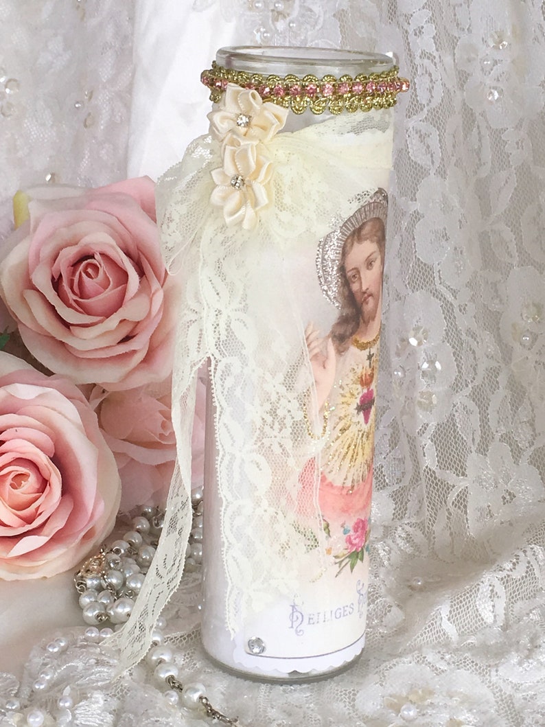 May include: A decorative candle with an image of Jesus, adorned with lace, ribbon, and floral accents. The candle features a gold and pink jeweled trim at the top and is surrounded by pink roses and pearls.