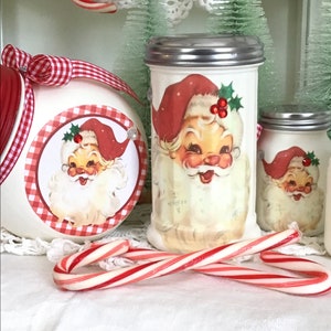May include: Two white ceramic sugar dispensers with red and white gingham trim and a Santa Claus image. The dispensers have silver lids. A candy cane is in the foreground.