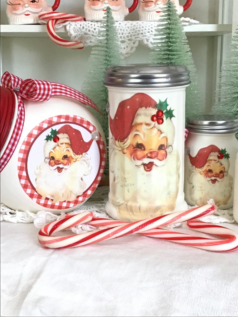 May include: Two white ceramic sugar dispensers with red and white gingham trim and a Santa Claus image. The dispensers have silver lids. A candy cane is in the foreground.