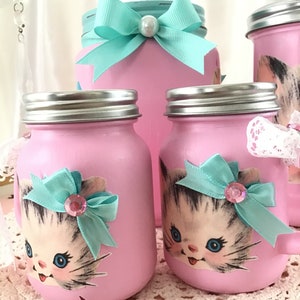 Puede incluir: Conjunto de cuatro tarros de vidrio rosa con tapas plateadas, decorados con caras de gatitos vintage y lazos turquesa. Los tarros son perfectos para guardar objetos peque&ntilde;os como az&uacute;car, t&eacute; o especias.