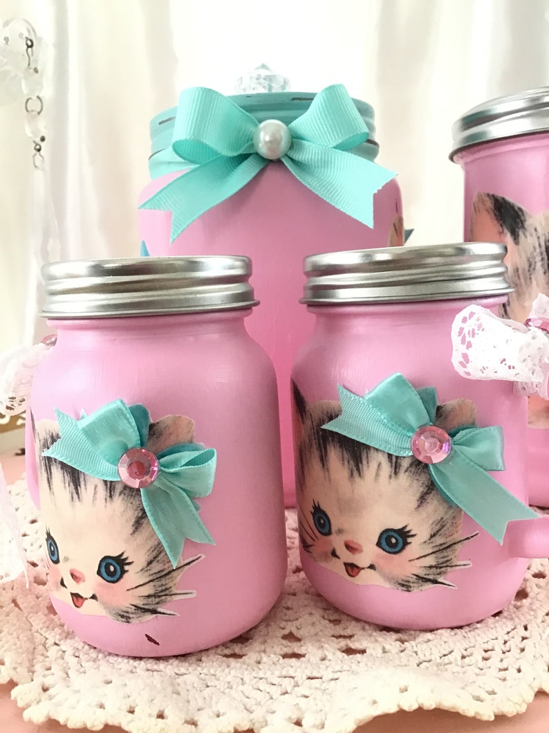 Puede incluir: Conjunto de cuatro tarros de vidrio rosa con tapas plateadas, decorados con caras de gatitos vintage y lazos turquesa. Los tarros son perfectos para guardar objetos peque&ntilde;os como az&uacute;car, t&eacute; o especias.