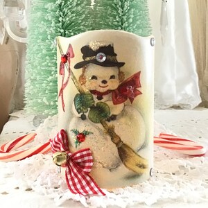 May include: A vintage-style snowman candle holder with a red and white gingham bow and a gold bell. The snowman is holding a broom and wearing a black hat with a red bow.