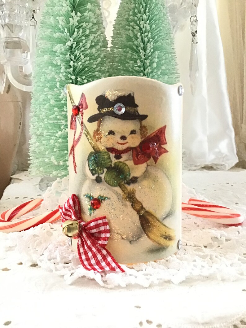May include: A vintage-style snowman candle holder with a red and white gingham bow and a gold bell. The snowman is holding a broom and wearing a black hat with a red bow.