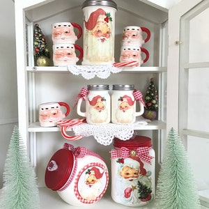 May include: White cabinet with glass doors and shelves filled with Christmas themed Santa Claus jars, mugs, and cookie jars. The jars are decorated with red and white gingham ribbon and a Santa Claus image. The mugs are white with a Santa Claus face and red trim. The cookie jars are red with a white gingham lid and a Santa Claus image.