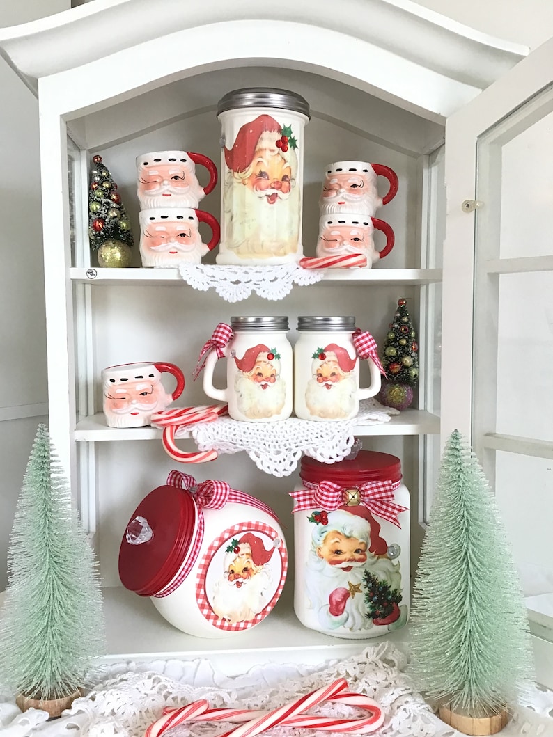 May include: White cabinet with glass doors and shelves filled with Christmas themed Santa Claus jars, mugs, and cookie jars. The jars are decorated with red and white gingham ribbon and a Santa Claus image. The mugs are white with a Santa Claus face and red trim. The cookie jars are red with a white gingham lid and a Santa Claus image.