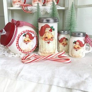 May include: White ceramic Santa Claus themed sugar and spice jars with red and white gingham ribbon and candy canes. The jars have red lids and are decorated with a Santa Claus image.