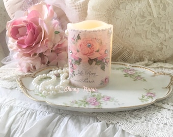 May include: A white pillar candle with a floral design and the text "Le Reve, Maison de Paris, Fanny Nippin". The candle is sitting on a white tray with a floral design and a pearl necklace.
