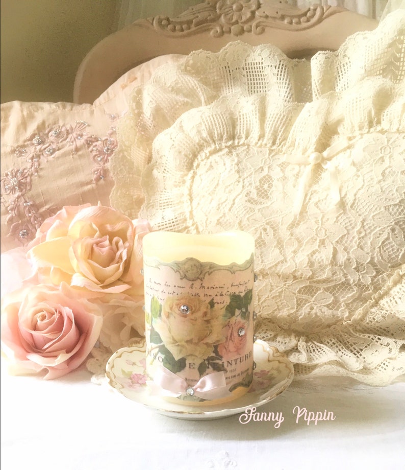 May include: A white pillar candle with a floral design and pink ribbon. The candle is decorated with rhinestones and sits on a small white saucer with a floral design. The candle is surrounded by pink and white roses.