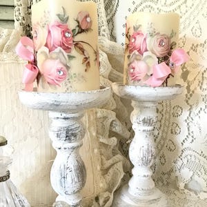 May include: Two pillar candles with rose floral patterns and pink ribbon embellishments. They are placed on distressed white wooden candle holders, giving a vintage feel. The candles are cream-colored.