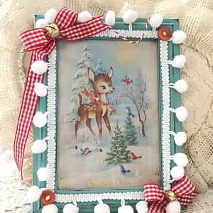 Puede incluir: Un marco navide&ntilde;o con una impresi&oacute;n de un ciervo en un paisaje nevado, adornado con cinta de cuadros rojos y blancos, campanas y pompones blancos. El marco est&aacute; pintado de color turquesa y tiene un ribete de encaje blanco. La impresi&oacute;n dice "Christmas Cheer".