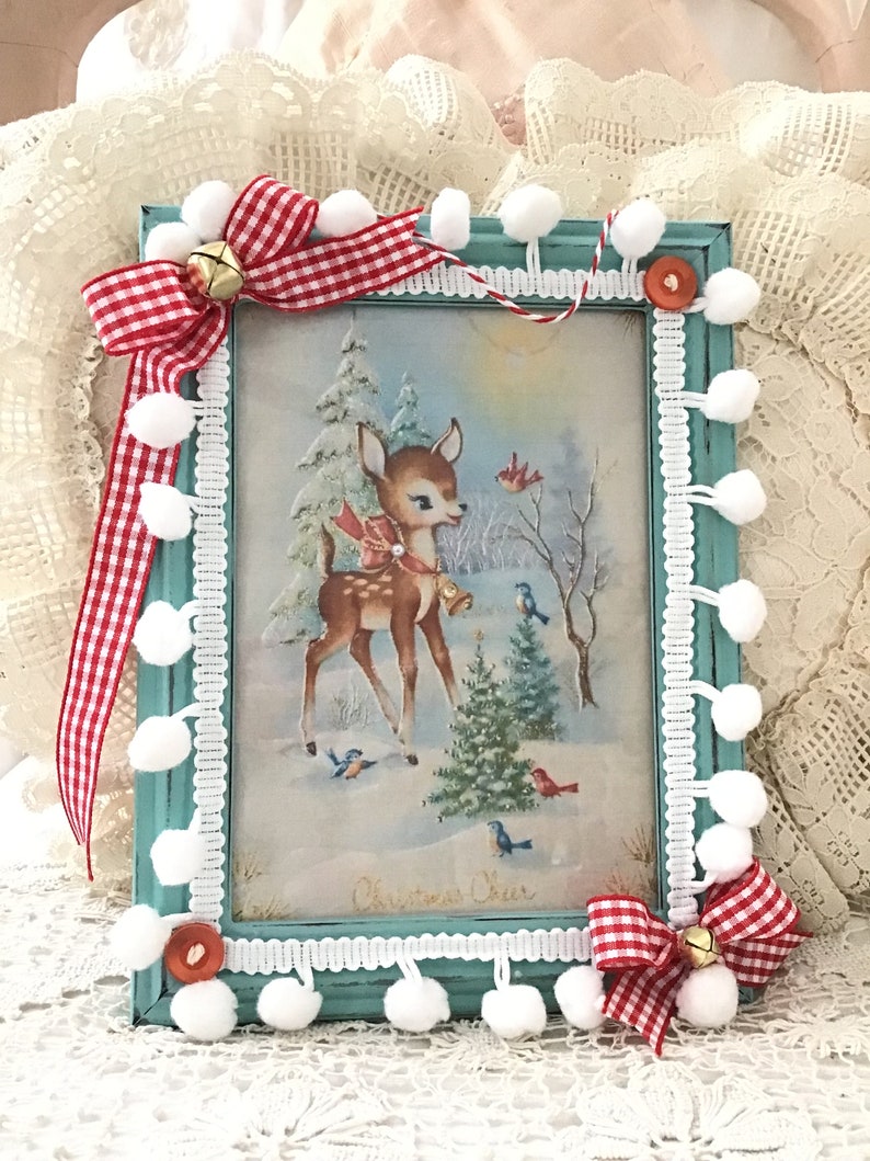 Puede incluir: Un marco navide&ntilde;o con una impresi&oacute;n de un ciervo en un paisaje nevado, adornado con cinta de cuadros rojos y blancos, campanas y pompones blancos. El marco est&aacute; pintado de color turquesa y tiene un ribete de encaje blanco. La impresi&oacute;n dice "Christmas Cheer".