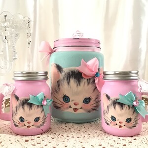 Puede incluir: Conjunto de tres tarros de cristal decorados con una imagen vintage de un gatito. Los tarros est&aacute;n pintados en rosa y azul con un lazo de cinta rosa y azul. El tarro m&aacute;s grande tiene una tapa con un pomo de cristal.
