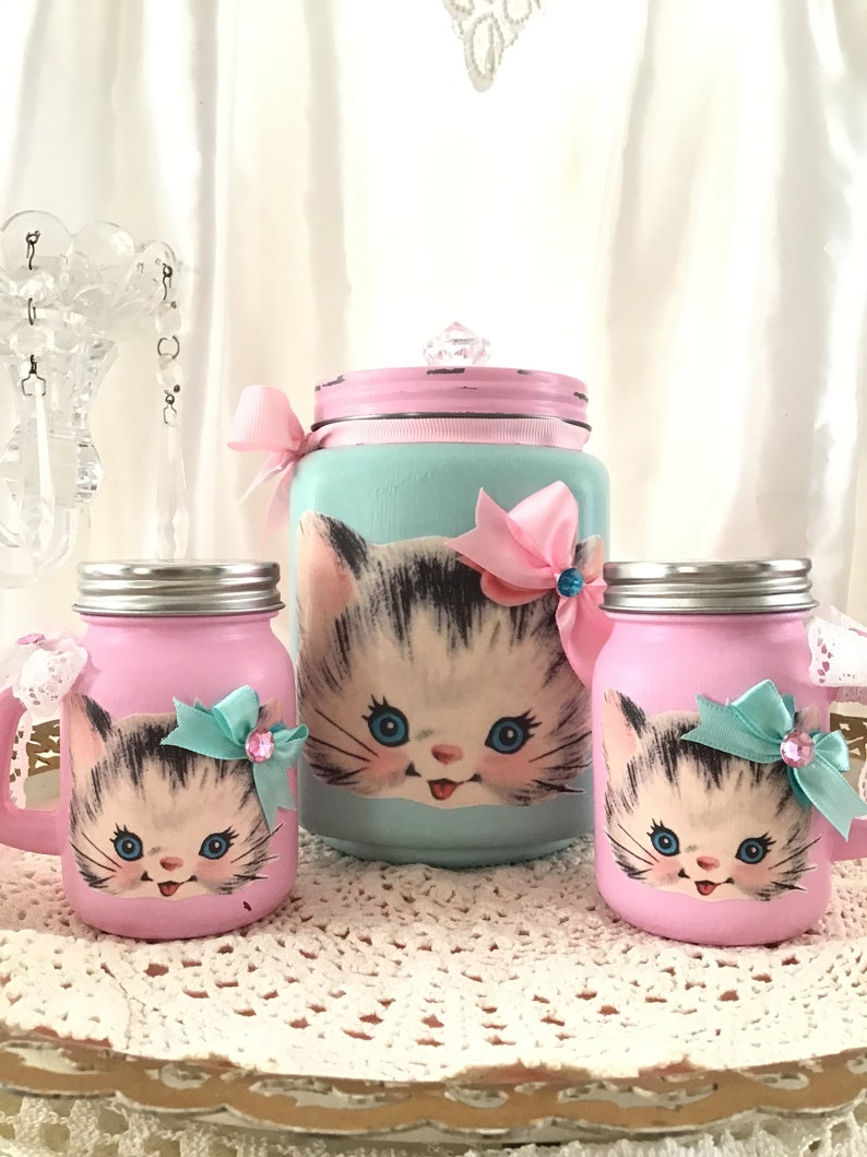 Puede incluir: Conjunto de tres tarros de cristal decorados con una imagen vintage de un gatito. Los tarros est&aacute;n pintados en rosa y azul con un lazo de cinta rosa y azul. El tarro m&aacute;s grande tiene una tapa con un pomo de cristal.