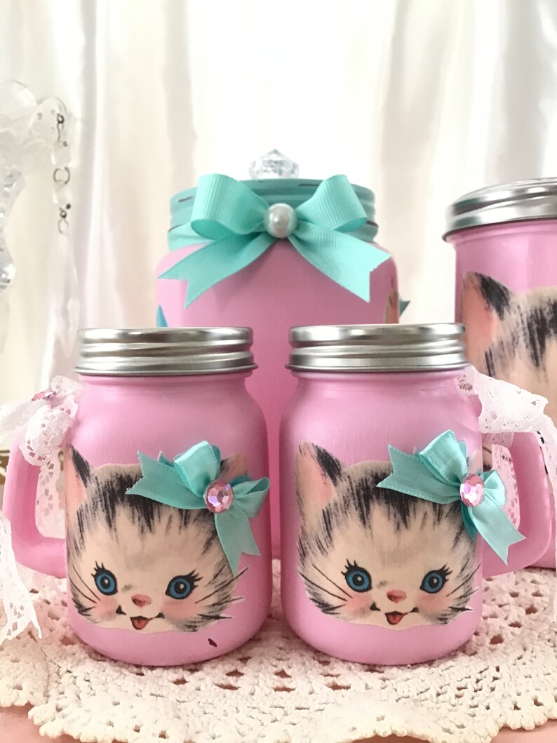 Puede incluir: Conjunto de tres tarros de cristal rosa con un dise&ntilde;o de gatito. Los tarros tienen una cinta turquesa y un adorno de strass. Los tarros son perfectos para guardar objetos peque&ntilde;os o para usar como elemento decorativo.