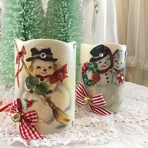 May include: Two white candle holders with a snowman design. The candle holders are decorated with red and white gingham ribbon and bells.