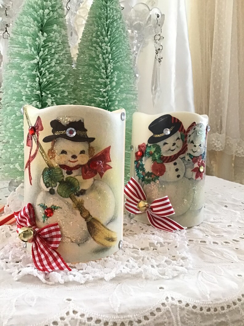 May include: Two white candle holders with a snowman design. The candle holders are decorated with red and white gingham ribbon and bells.