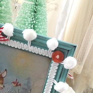 Puede incluir: Un marco de fotos turquesa con una escena invernal con un ciervo y p&aacute;jaros. El marco est&aacute; decorado con pompones blancos, ribete de encaje blanco y un bot&oacute;n rojo. Dos peque&ntilde;os &aacute;rboles de Navidad verdes est&aacute;n en el fondo.