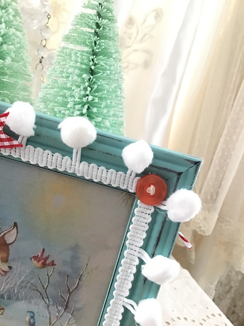 Puede incluir: Un marco de fotos turquesa con una escena invernal con un ciervo y p&aacute;jaros. El marco est&aacute; decorado con pompones blancos, ribete de encaje blanco y un bot&oacute;n rojo. Dos peque&ntilde;os &aacute;rboles de Navidad verdes est&aacute;n en el fondo.