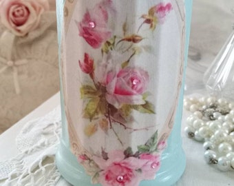 Puede incluir: Un tarro de cerámica azul claro con un diseño floral con rosas rosas y hojas verdes. El tarro tiene un fondo blanco y un borde decorativo. El tarro está decorado con pequeños diamantes de imitación transparentes.