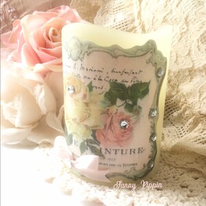 May include: A white pillar candle with a vintage floral design and rhinestones. The design features roses and a text label that reads "E. INTURE 1902" and "INVENTS SANS LES DECORER".