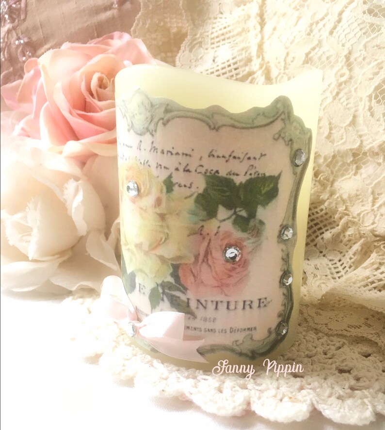 May include: A white pillar candle with a vintage floral design and rhinestones. The design features roses and a text label that reads "E. INTURE 1902" and "INVENTS SANS LES DECORER".