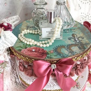 May include: A decorative round box with a teal floral design, adorned with a pink bow and pearl necklace. The box holds perfume bottles and a small jar. The box is trimmed with gold and pink tassels. The perfume bottle reads "Macaron Rose".
