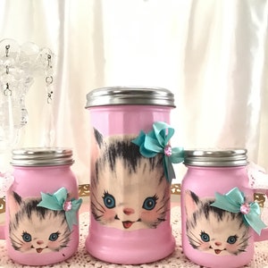 Puede incluir: Conjunto de tres tarros de vidrio rosa con un dise&ntilde;o de gatito vintage. Cada tarro tiene una tapa plateada y un lazo turquesa con un acento de strass. Los tarros est&aacute;n sobre un encaje blanco.