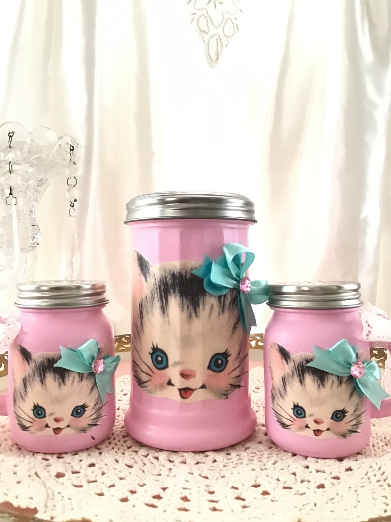 Puede incluir: Conjunto de tres tarros de vidrio rosa con un dise&ntilde;o de gatito vintage. Cada tarro tiene una tapa plateada y un lazo turquesa con un acento de strass. Los tarros est&aacute;n sobre un encaje blanco.