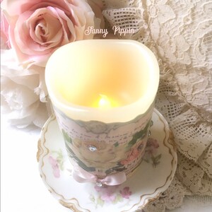 May include: A white candle with a floral design and a pink ribbon sits on a white saucer with a floral pattern. The candle is lit and the flame is visible. The text "Sanny Pippin" is visible on the candle.
