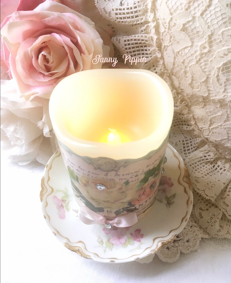 May include: A white candle with a floral design and a pink ribbon sits on a white saucer with a floral pattern. The candle is lit and the flame is visible. The text "Sanny Pippin" is visible on the candle.