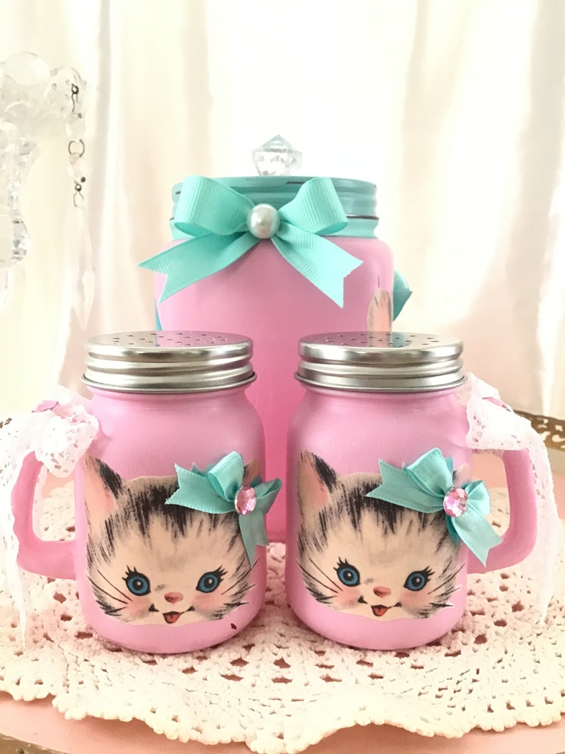 Puede incluir: Conjunto de tres tarros de vidrio rosa con caras de gatitos y lazos turquesa. Los tarros tienen tapas plateadas y est&aacute;n decorados con una cara de gatito de estilo vintage. El tarro m&aacute;s grande tiene un lazo turquesa y los tarros m&aacute;s peque&ntilde;os tienen un borde de encaje blanco.
