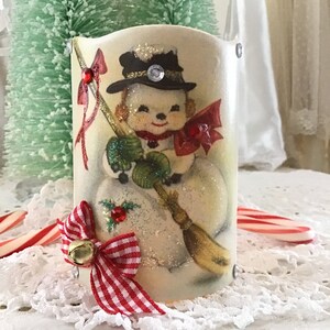 May include: A white ceramic candle holder with a vintage illustration of a snowman wearing a black hat and red scarf. The holder is decorated with a red and white gingham bow and a gold bell.