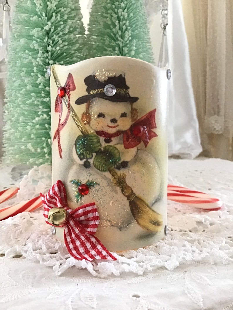 May include: A white ceramic candle holder with a vintage illustration of a snowman wearing a black hat and red scarf. The holder is decorated with a red and white gingham bow and a gold bell.