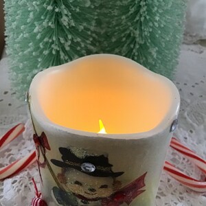 May include: A white ceramic candle with a snowman design and a flickering flame inside. The candle is decorated with red and gold accents and a single rhinestone.