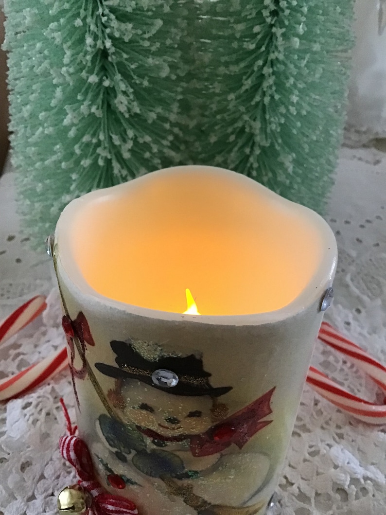 May include: A white ceramic candle with a snowman design and a flickering flame inside. The candle is decorated with red and gold accents and a single rhinestone.