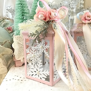 May include: A pink decorative lantern with snowflake cutouts, adorned with pink roses, pearls, and ribbons. The lantern is decorated with white snowflakes and sits on a white lace surface. The background includes small green Christmas trees.