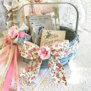 May include: A light blue woven basket filled with vintage books, including one titled "British Song Birds." The basket is adorned with a large floral bow and pink ribbons, with a backdrop of lace and fabric roses. The word "SHABBY" is visible on one of the books.